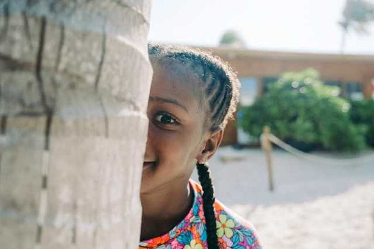 A Black child peers at the camera from behind a tree trunk