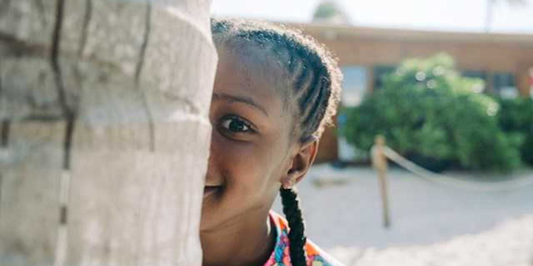 A Black child peers at the camera from behind a tree trunk