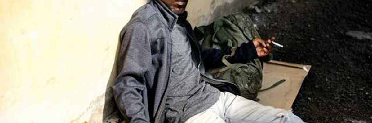 A Black homeless man sits on a piece of cardboard