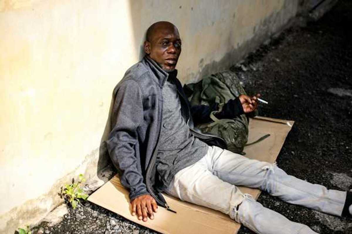 A Black homeless man sits on a piece of cardboard