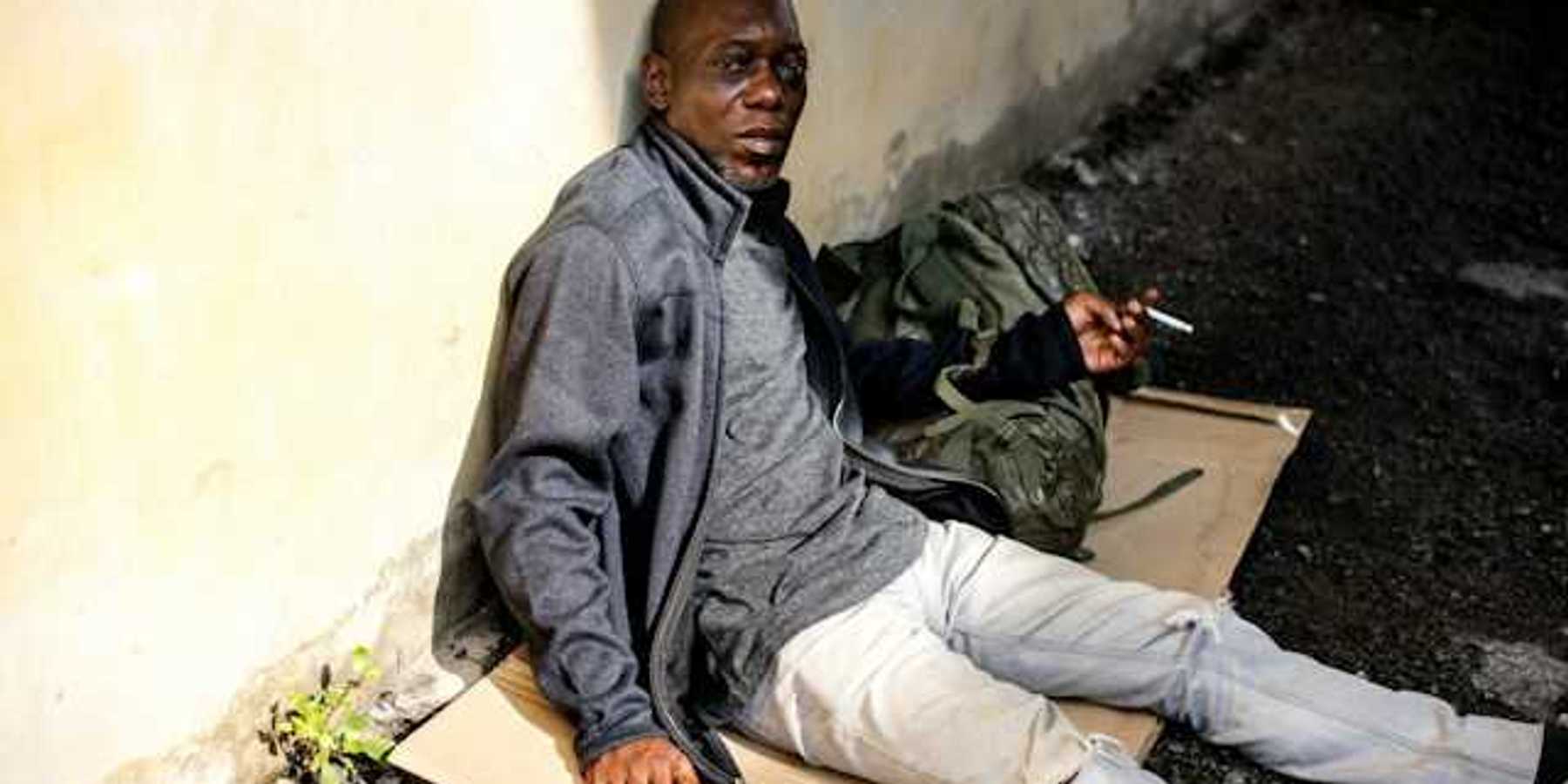 A Black homeless man sits on a piece of cardboard