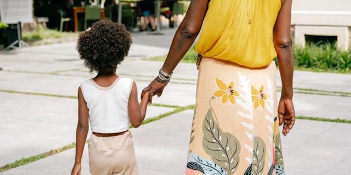 A black mother wearing a yellow top and floral print skirt holding a child's hand