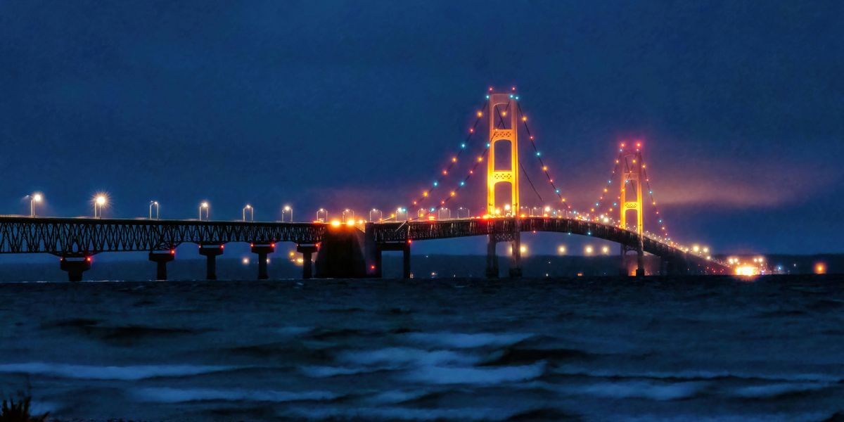a bridge that is lit up at night with wave breaking on a nearby lake shore in foreground.