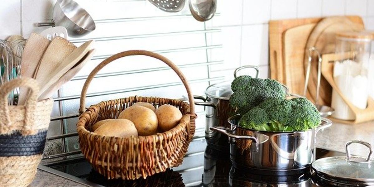 A closeup of an electric cooktop with pots of broccoli and a basket of potatoes on top of the range.