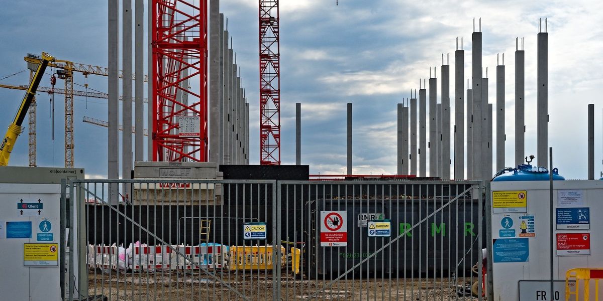 A construction site with cranes and concrete support structures behind a closed gate.