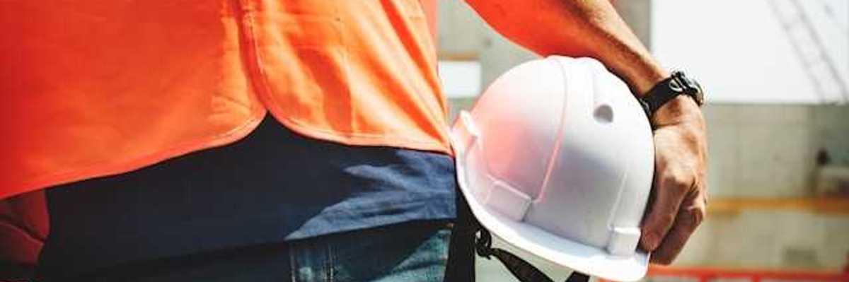 A construction worker in an orange vest carrying a white hard hat on a sunny day