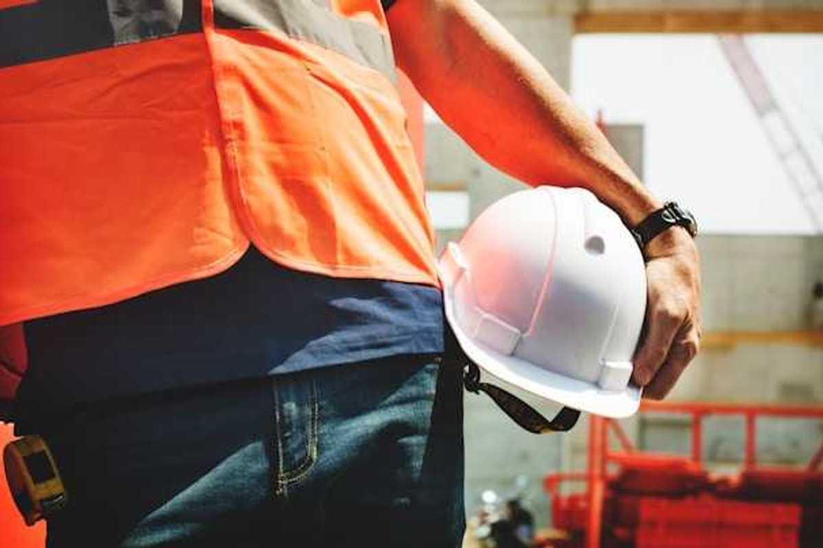 A construction worker in an orange vest carrying a white hard hat on a sunny day