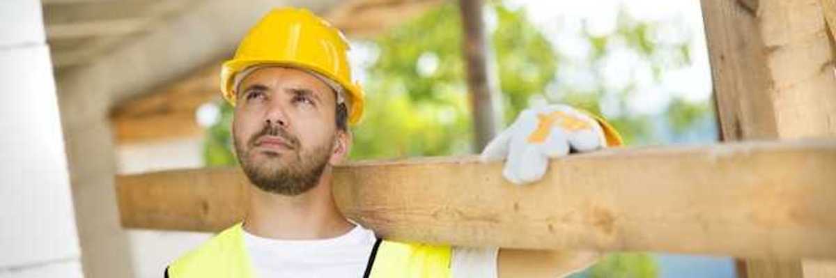 A construction worker wearing a white tshirt and a yellow safety vest carrying a piece of wood over his shoulder