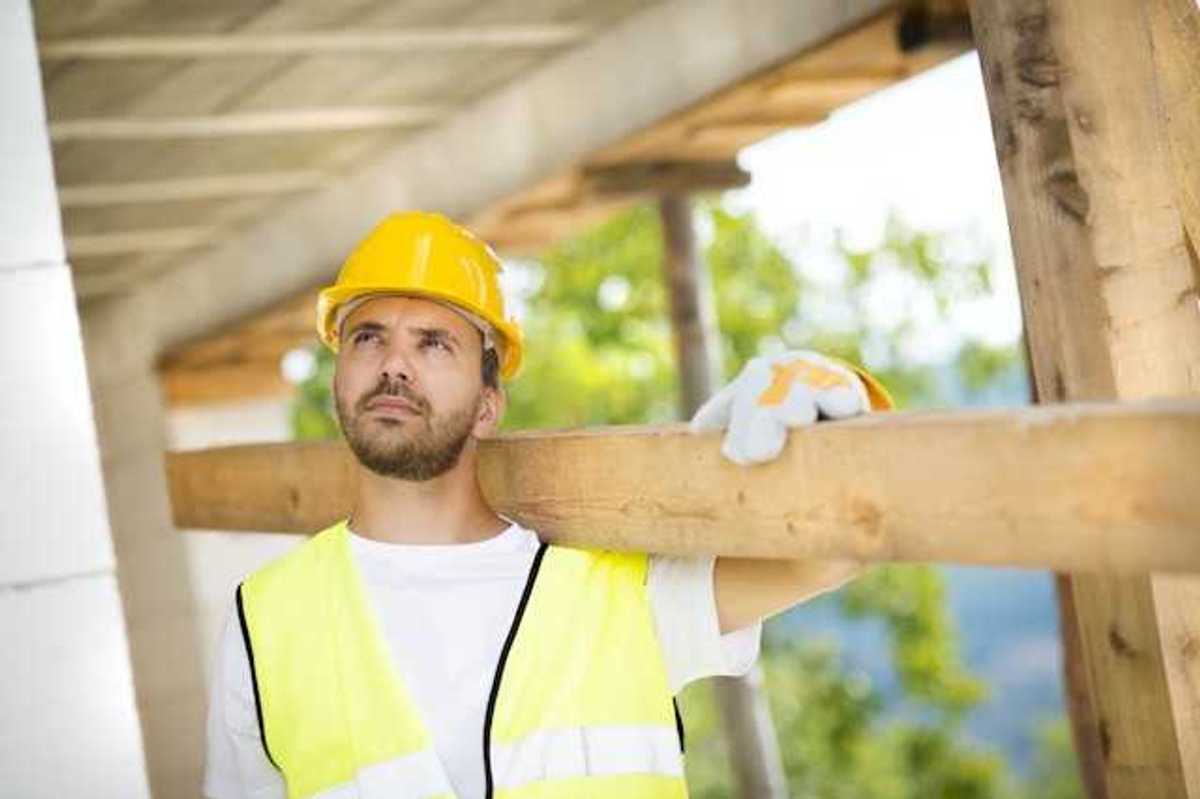 A construction worker wearing a white tshirt and a yellow safety vest carrying a piece of wood over his shoulder