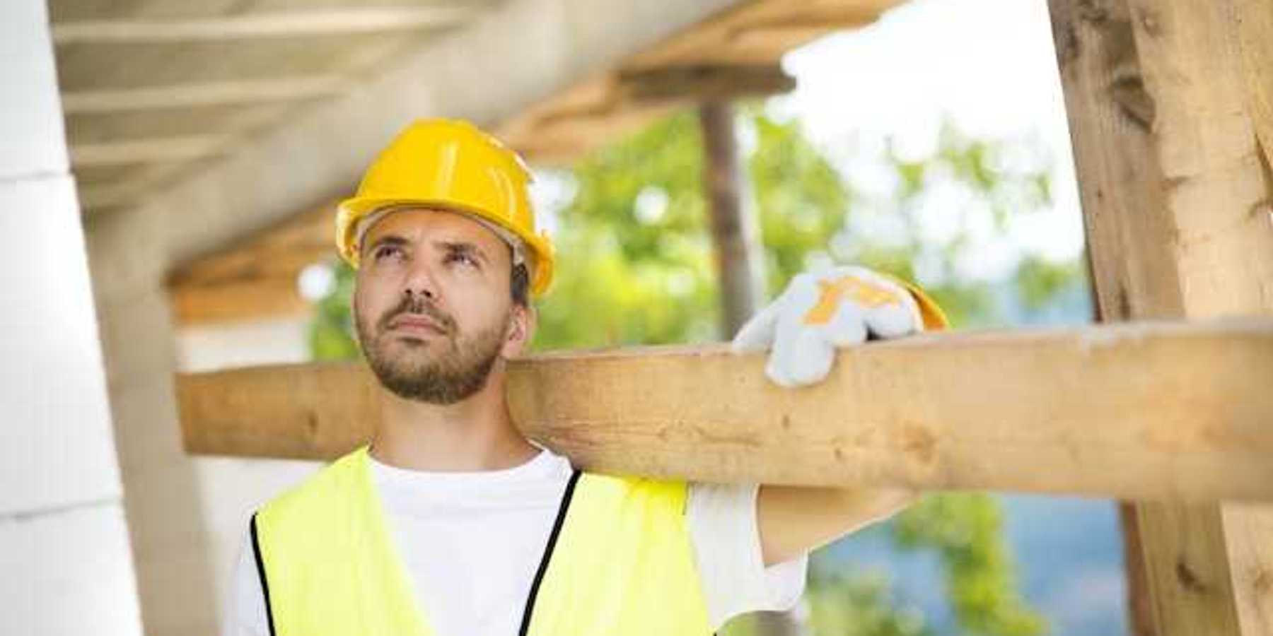 A construction worker wearing a white tshirt and a yellow safety vest carrying a piece of wood over his shoulder