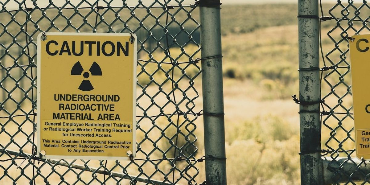 A cyclone fence gate locked with a chain and padlock displays a yellow caution sign about underground radioactive material.