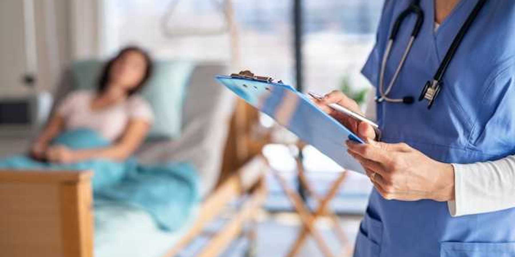 A doctor with a clipboard stands in a hospital room with a patient in a bed in the background