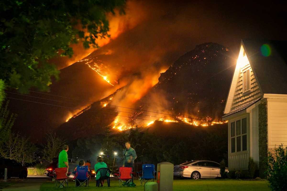 A family watches a nearby wildfire from their front lawn as flames cover a nearby mountainside in a nighttime setting