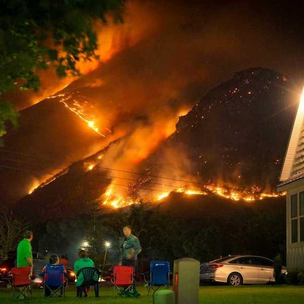 A family watches a nearby wildfire from their front lawn as flames cover a nearby mountainside in a nighttime setting