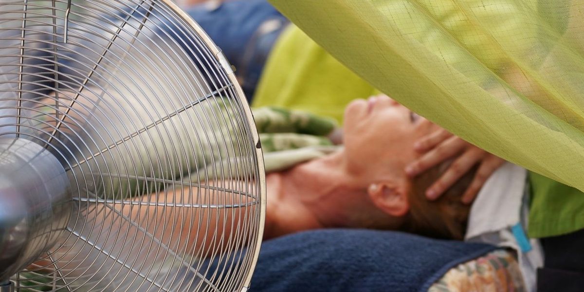 A fan blows on a woman laying down under a green mosquito net.