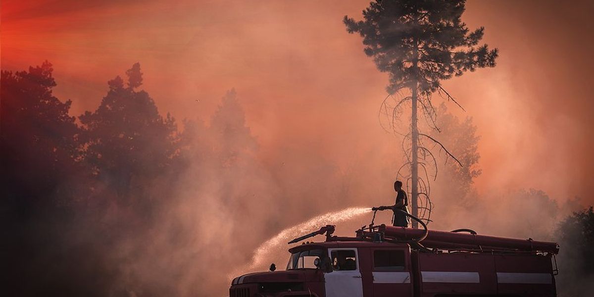 A firefighter stands on an old fire truck spraying water on grass with wildfire smoke in the air