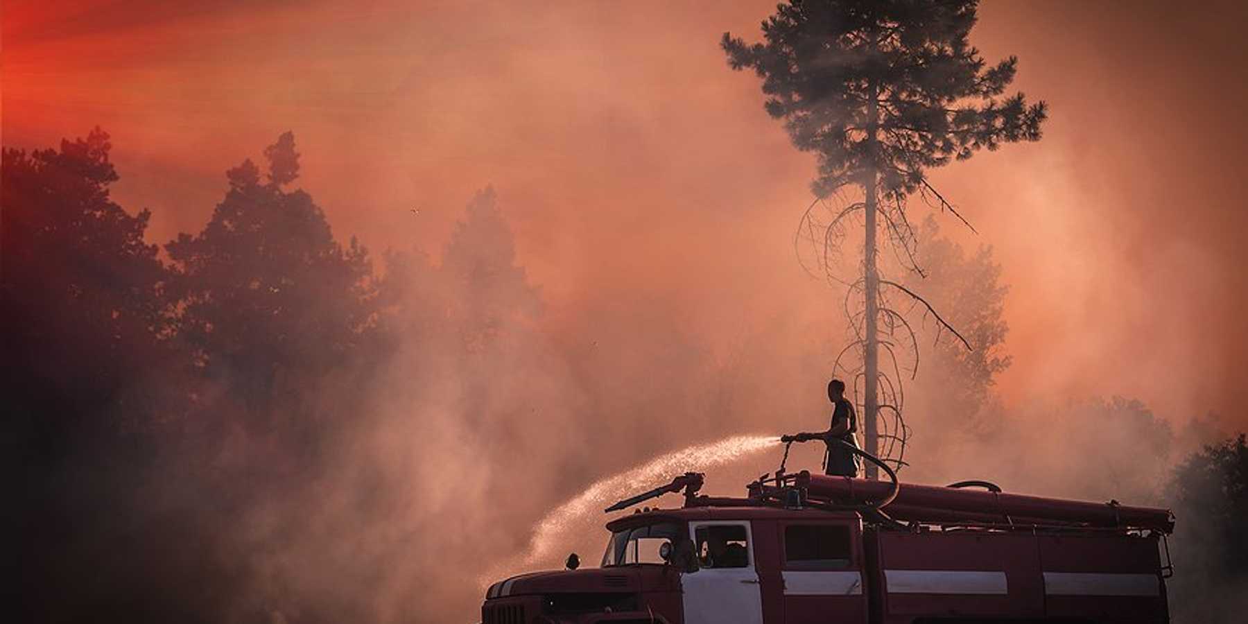 A firefighter stands on an old fire truck spraying water on grass with wildfire smoke in the air