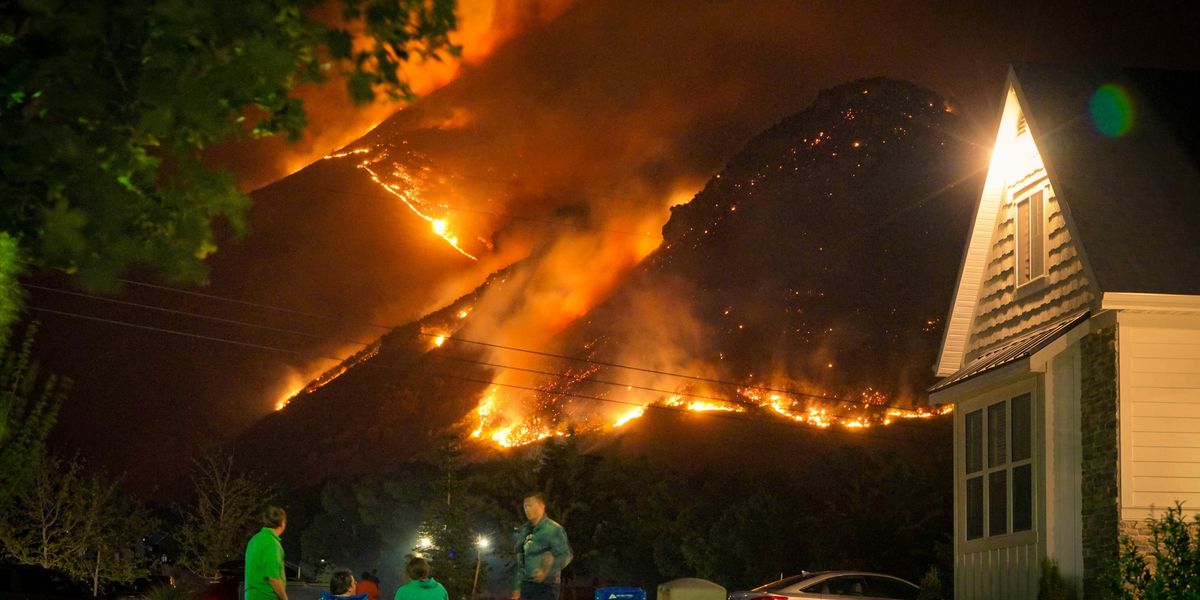 a group of people sitting in lawn chairs in a front yard at night, with a large wildfire illuminating a nearby hillside.