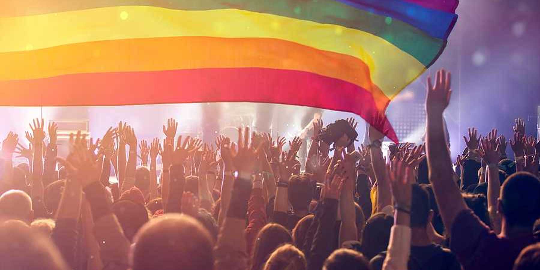 A group of people with their hands waving holding an LGBTQ flag in the air