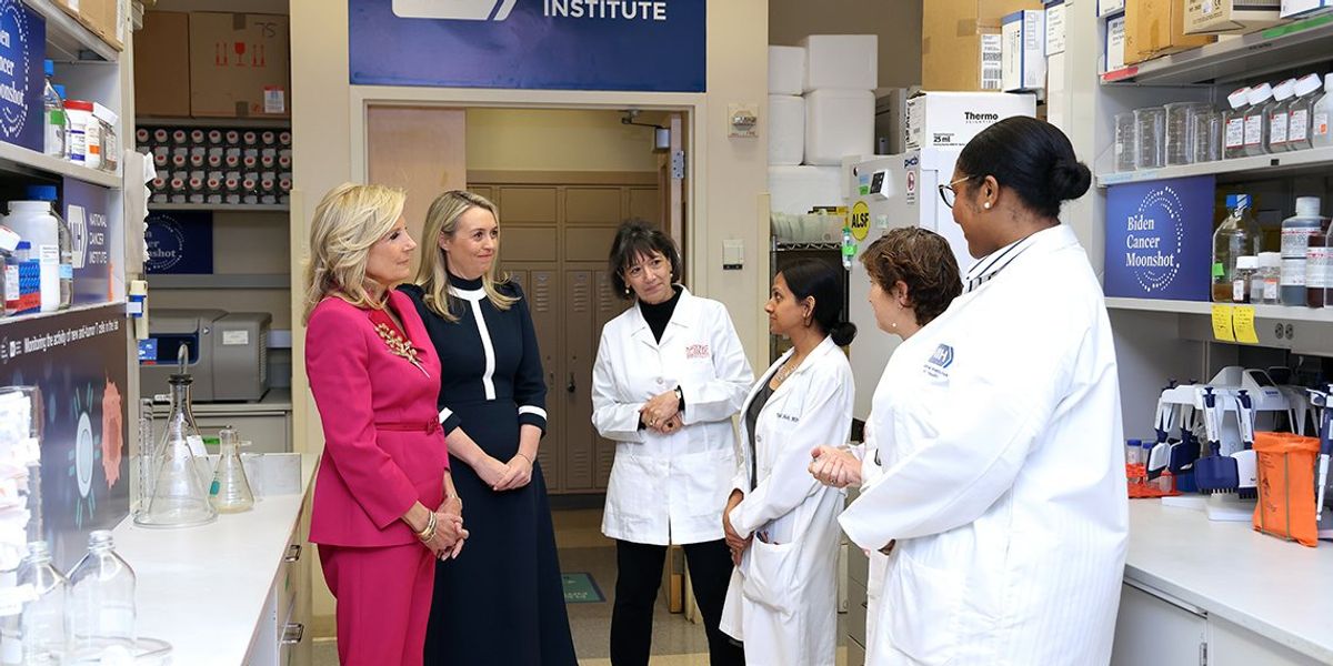 A group of scientists and women in suits converse in a lab of the National Cancer Institute.