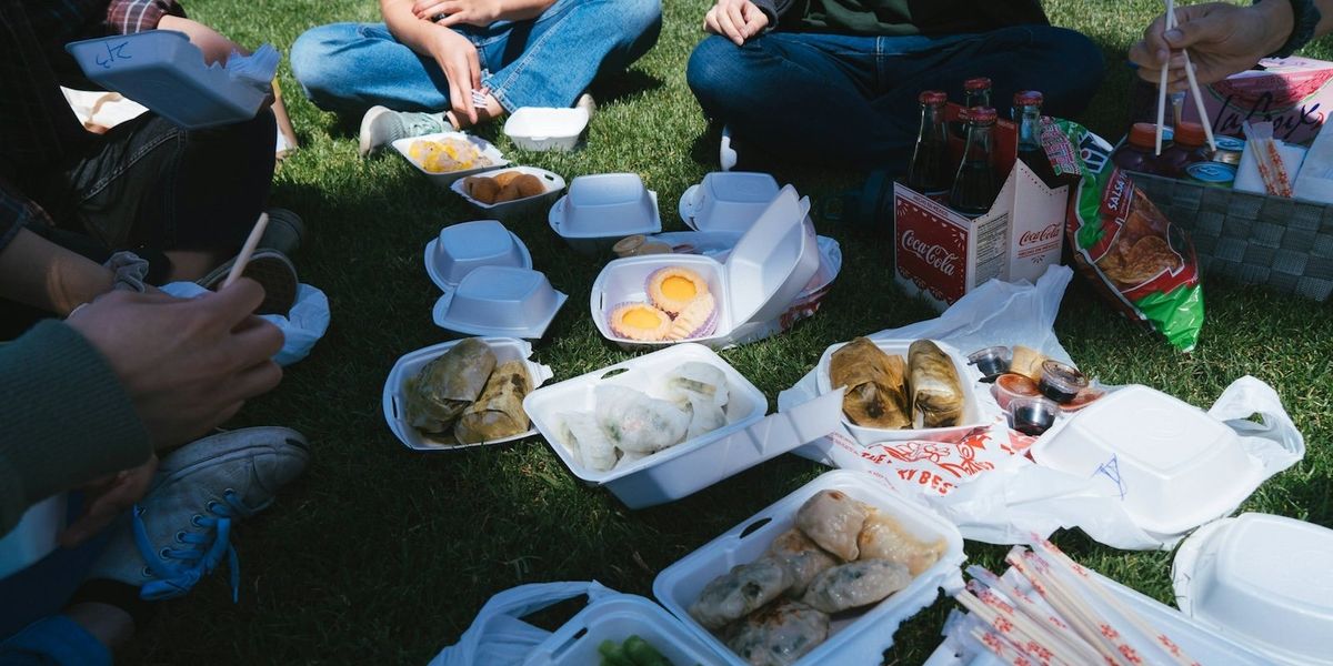 A group of teens sits on the grass eating fast food out of styrofoam containers with soda bottles nearby.