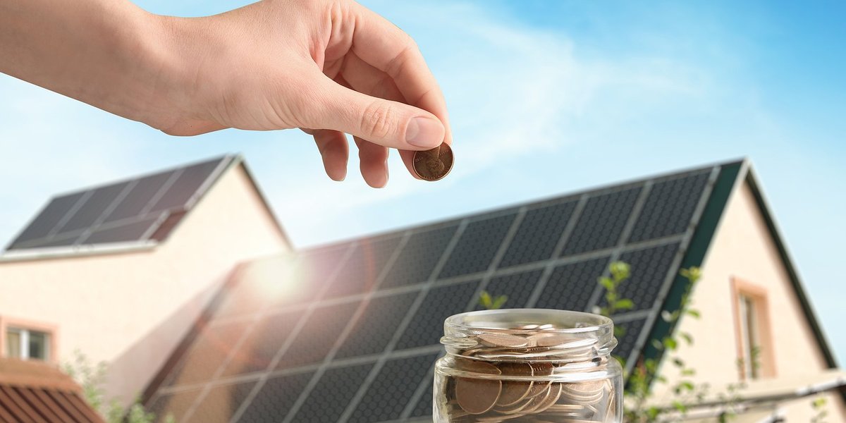 A hand places pennies in a glass jar with an image of a house roof covered in solar panels in background.