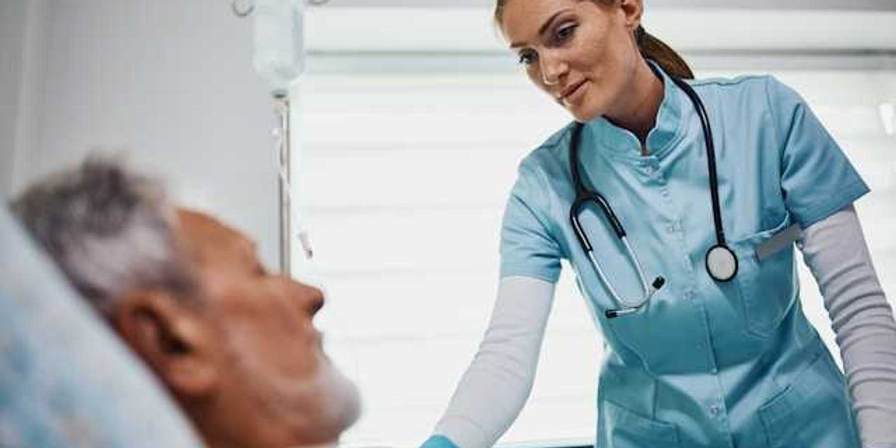 A health care professional wearing scrubs and a stethoscope leaning over an older patient in a bed