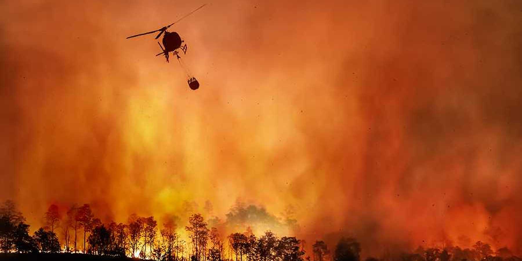 A helicopter dropping water onto a wildfire with burning trees in the background