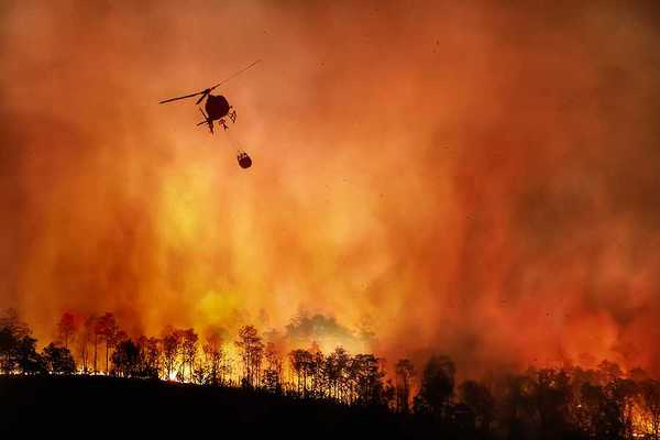 A helicopter dropping water onto a wildfire with burning trees in the background