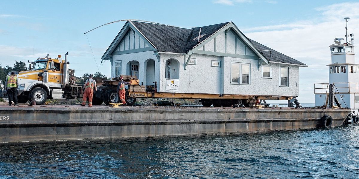 A house is loaded onto a truck at a dock next to a body of water.