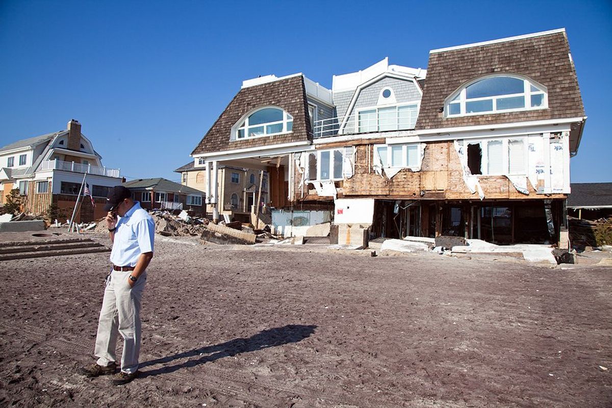 A hurricane damaged house with a man talking on the phone in front of it.