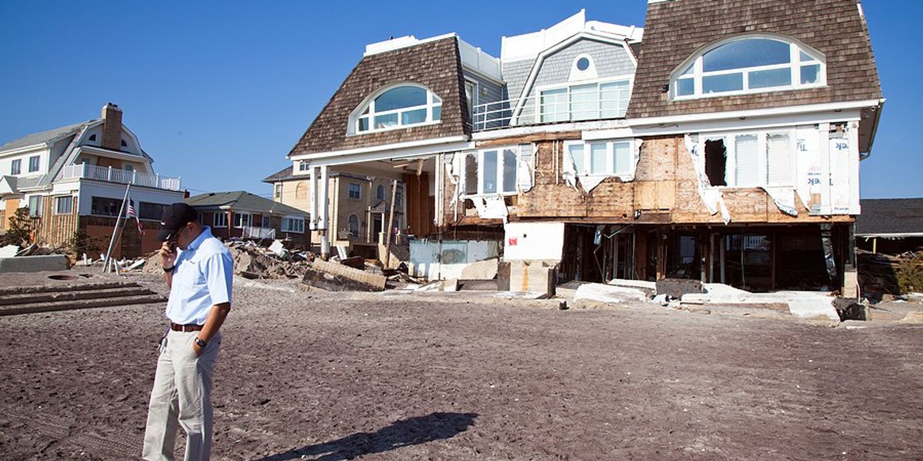 A hurricane damaged house with a man talking on the phone in front of it.