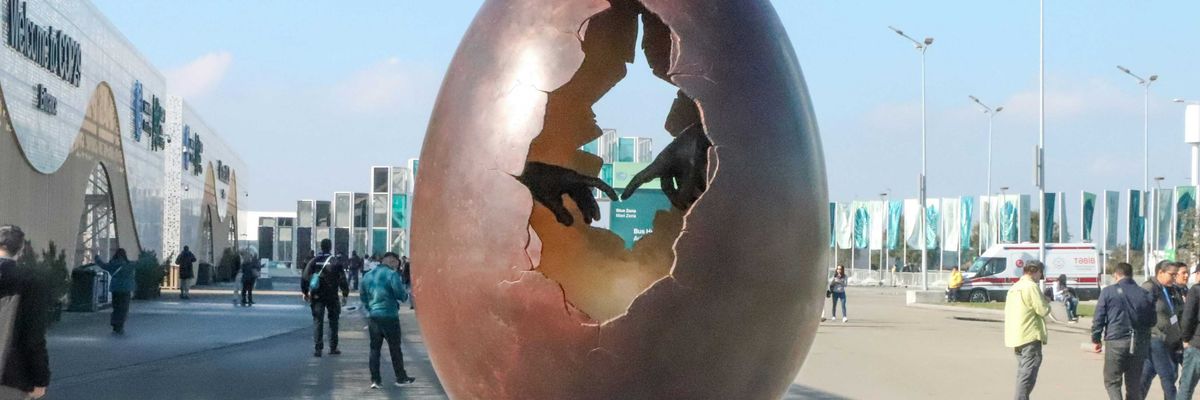 A large egg sculpture with a hole in the middle showing two hands with fingers touching, sitting on top of a metal pole.