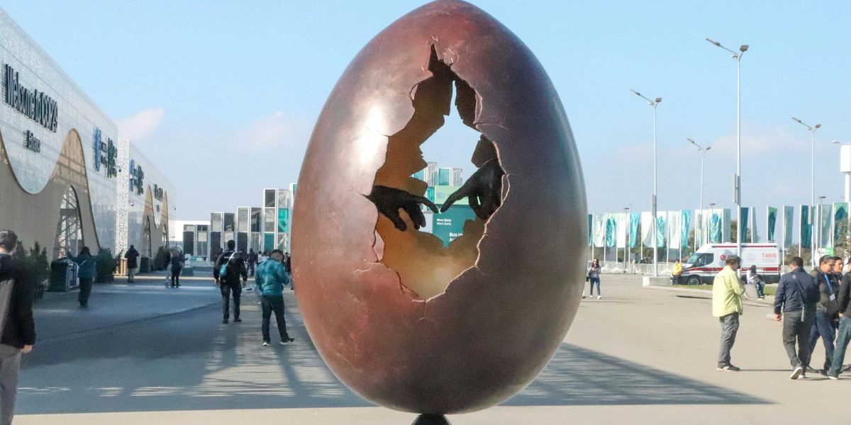 A large egg sculpture with a hole in the middle showing two hands with fingers touching, sitting on top of a metal pole.