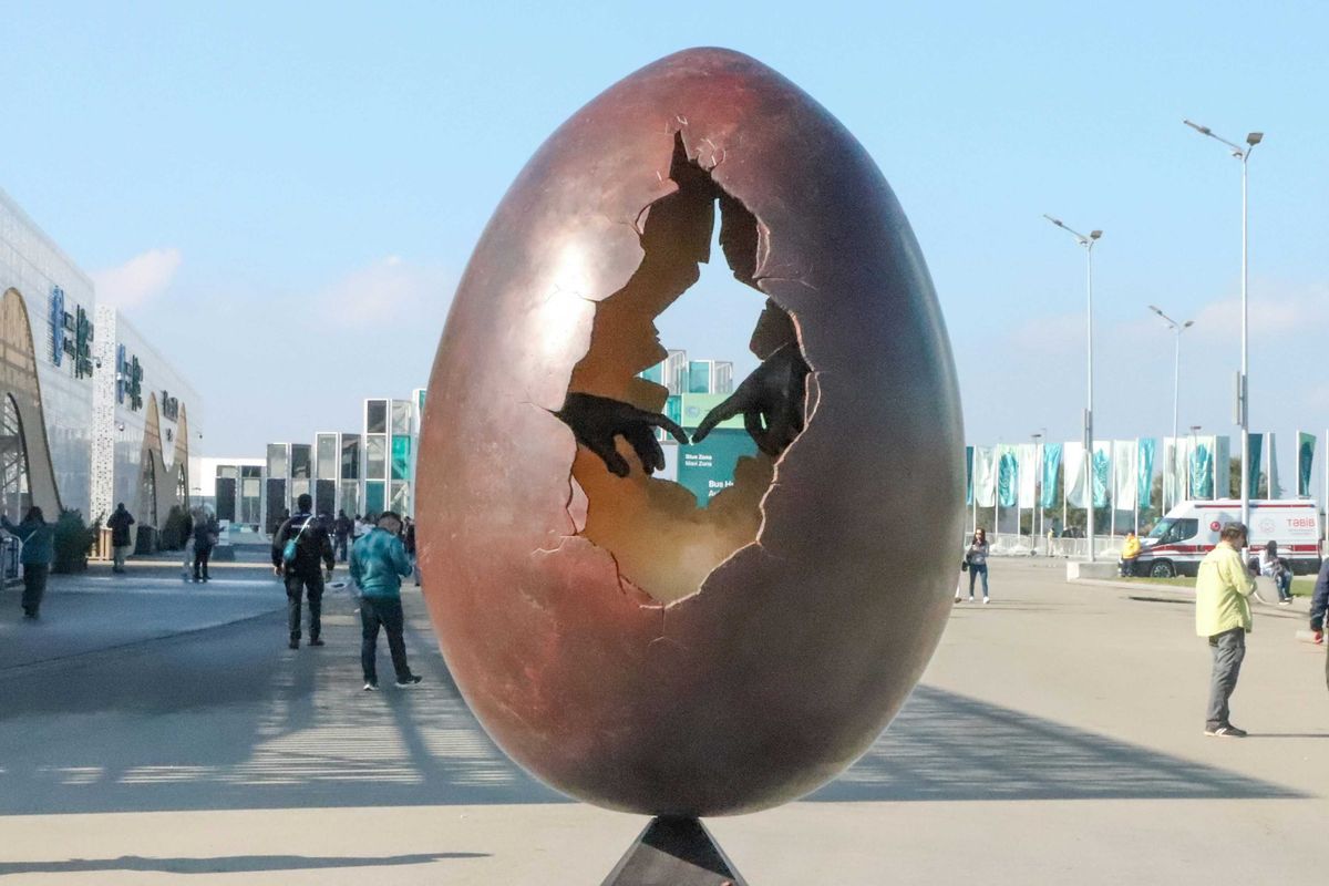 A large egg sculpture with a hole in the middle showing two hands with fingers touching, sitting on top of a metal pole.