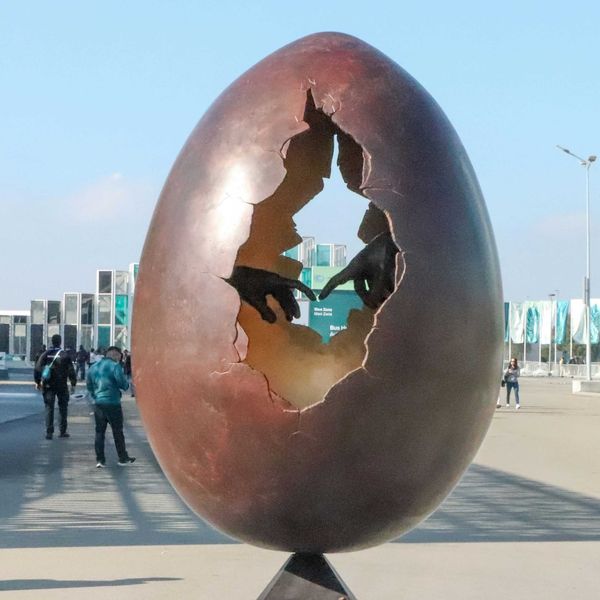 A large egg sculpture with a hole in the middle showing two hands with fingers touching, sitting on top of a metal pole.