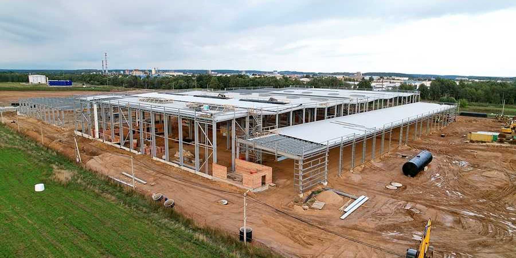 A large warehouse under construction with green fields and a town in the distance