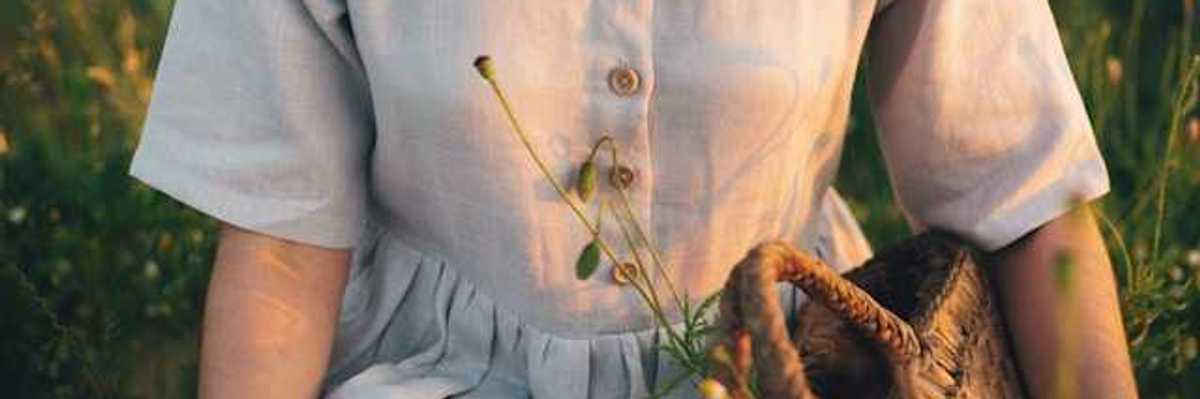 A linen dress on a woman in a field of wildflowers; she is holding a woven basket with plants inside