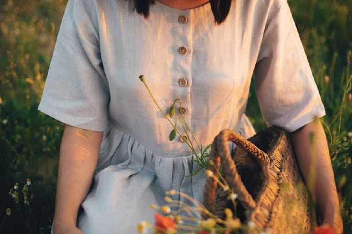 A linen dress on a woman in a field of wildflowers; she is holding a woven basket with plants inside