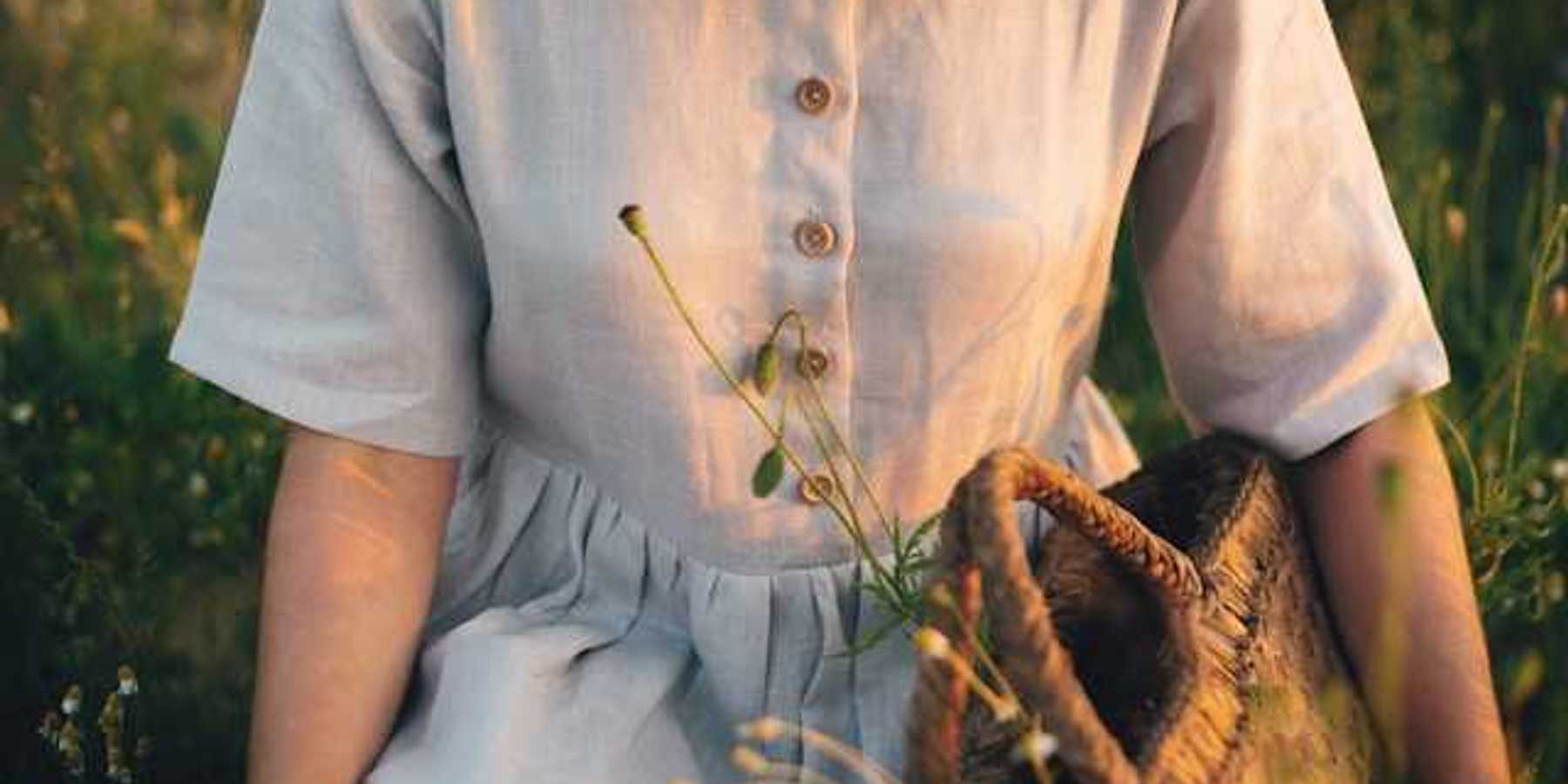 A linen dress on a woman in a field of wildflowers; she is holding a woven basket with plants inside