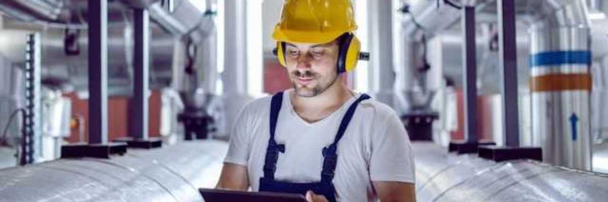 A maintenance worker looking at a tablet while standing next to giant silver pipes