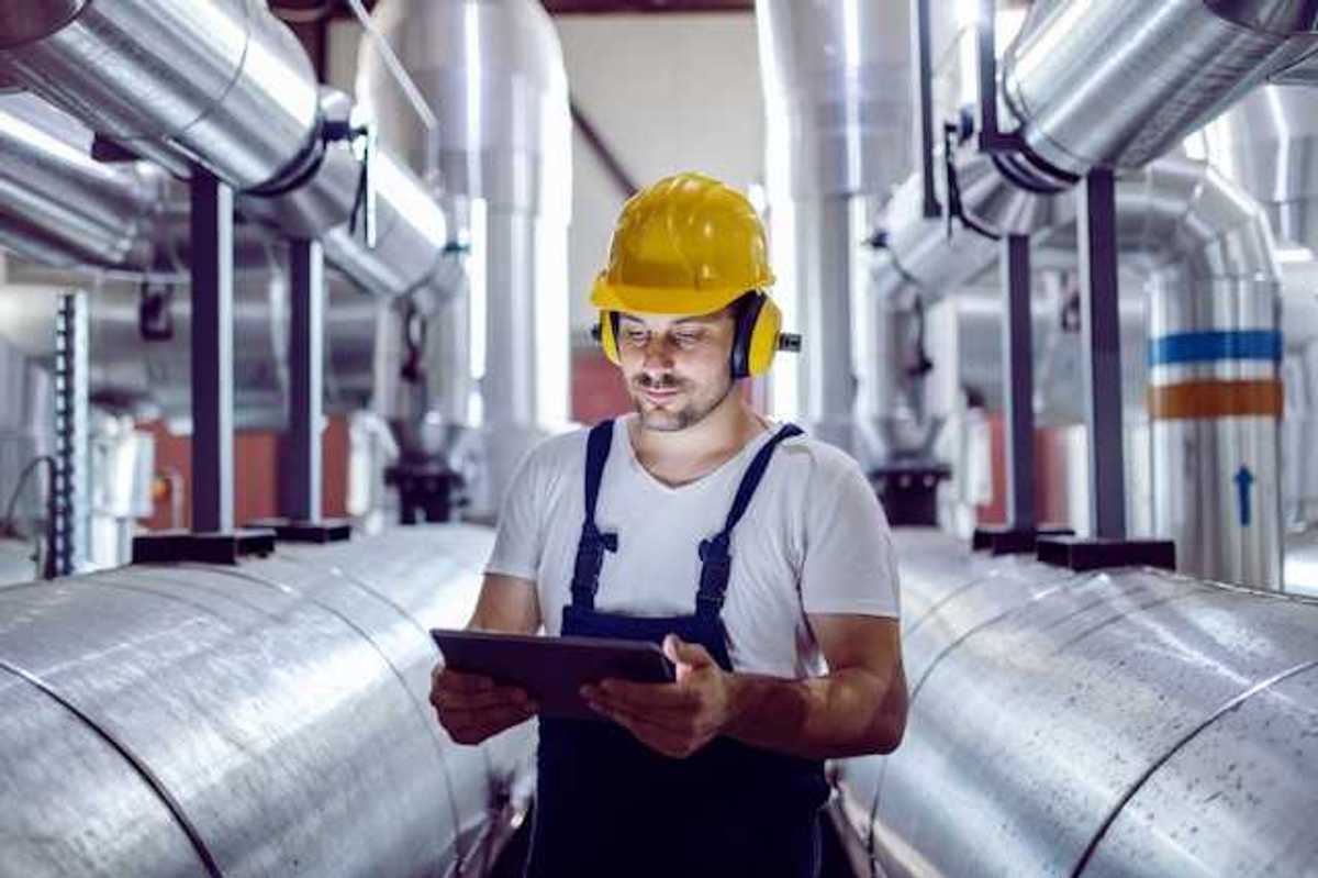 A maintenance worker looking at a tablet while standing next to giant silver pipes