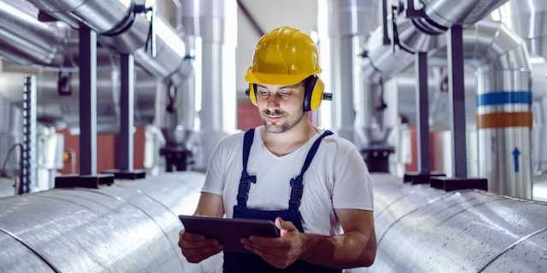 A maintenance worker looking at a tablet while standing next to giant silver pipes