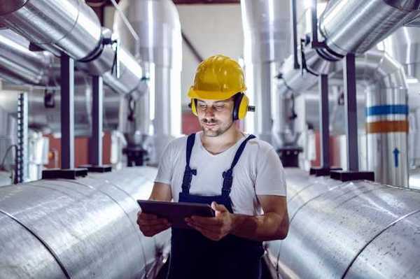 A maintenance worker looking at a tablet while standing next to giant silver pipes