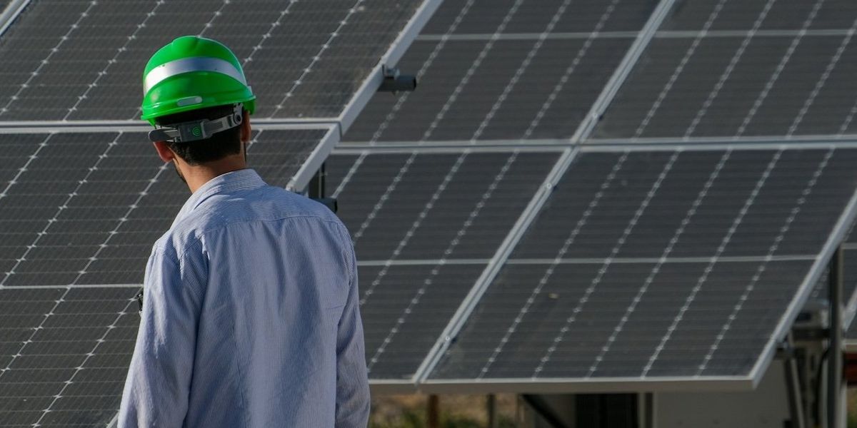 A man in a hard hat looks at solar panels in a golden landscape.