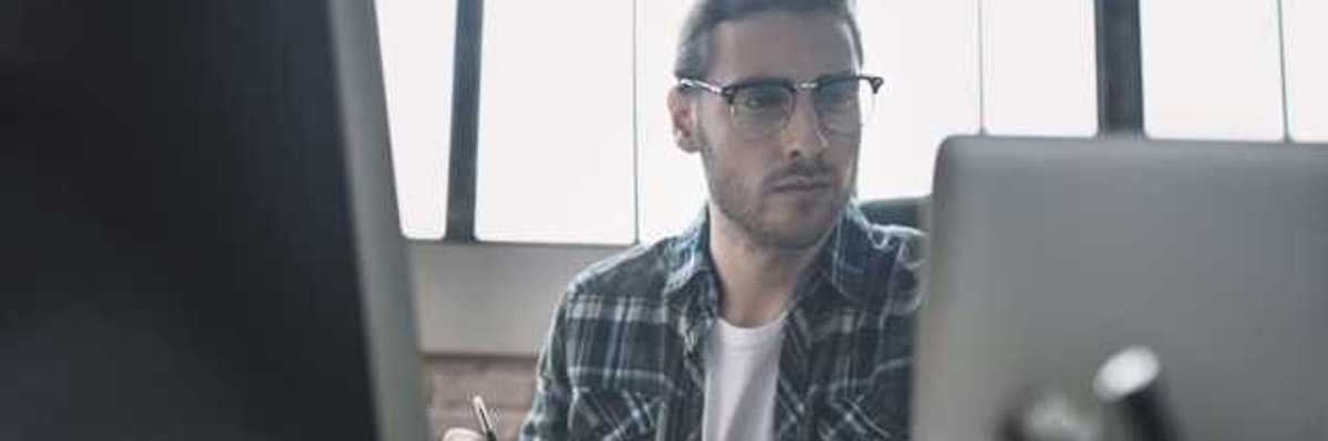 A man in a plaid shirt sitting at a desk in front of a laptop and monitor