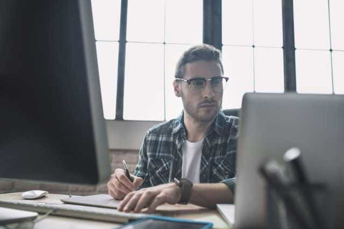 A man in a plaid shirt sitting at a desk in front of a laptop and monitor