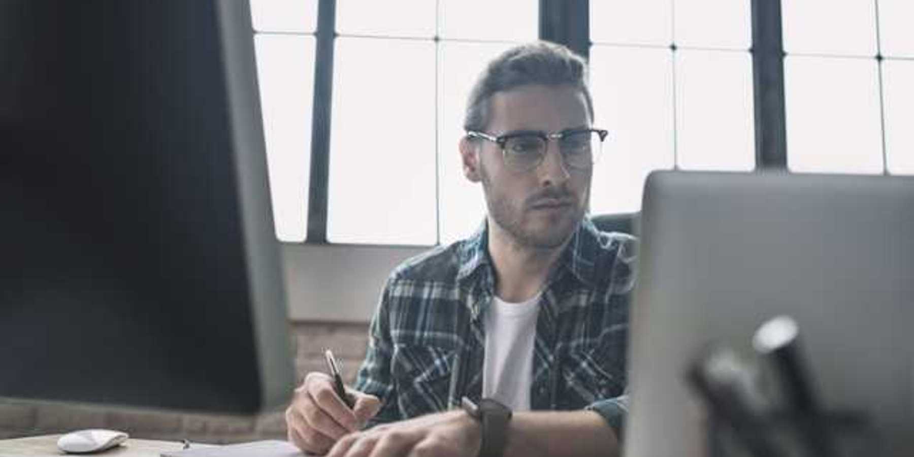 A man in a plaid shirt sitting at a desk in front of a laptop and monitor