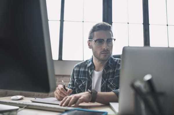 A man in a plaid shirt sitting at a desk in front of a laptop and monitor
