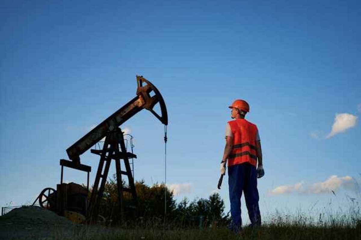 A man in an orange vest looking up at an oil pump jack